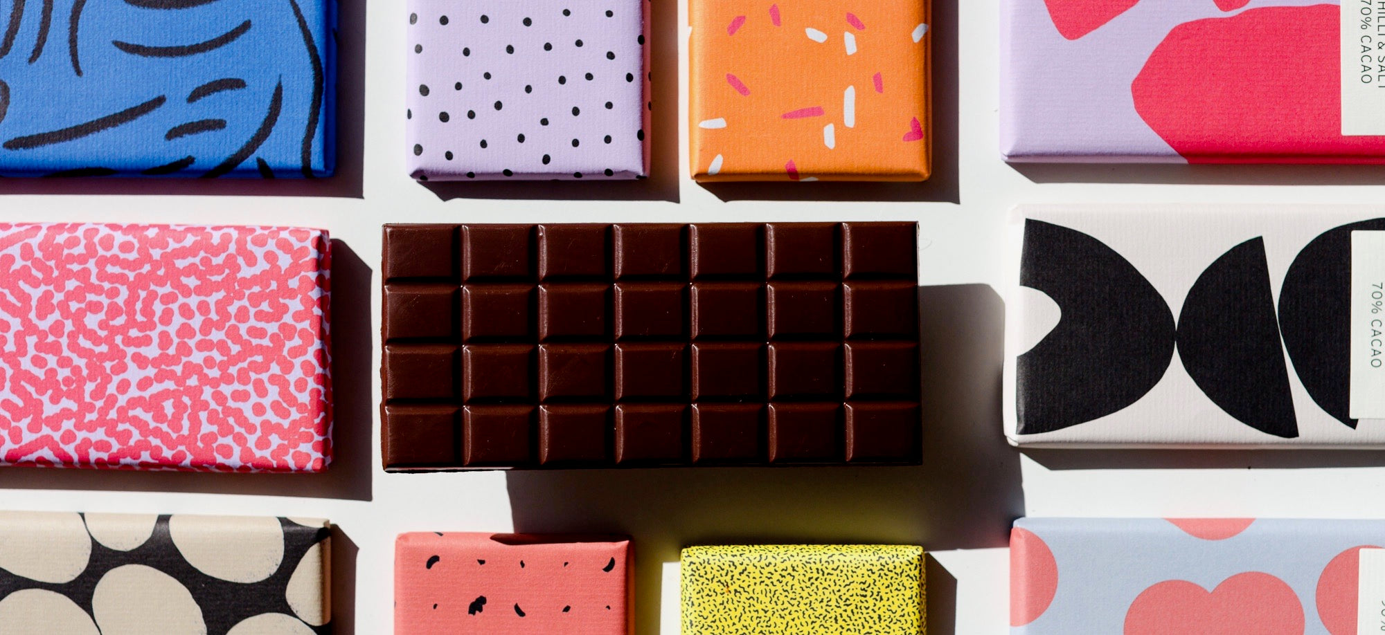 Overhead flat lay of Bahen & Co bean to bar chocolate bars in hand-designed patterned wrappers with one unwrapped dark chocolate bar, a Margaret River WA maker featured in No Matter What Gift Box corporate gift boxes